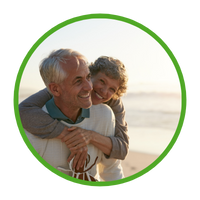 Older woman hugging older man from behind, both grinning and having fun while walking along beach.