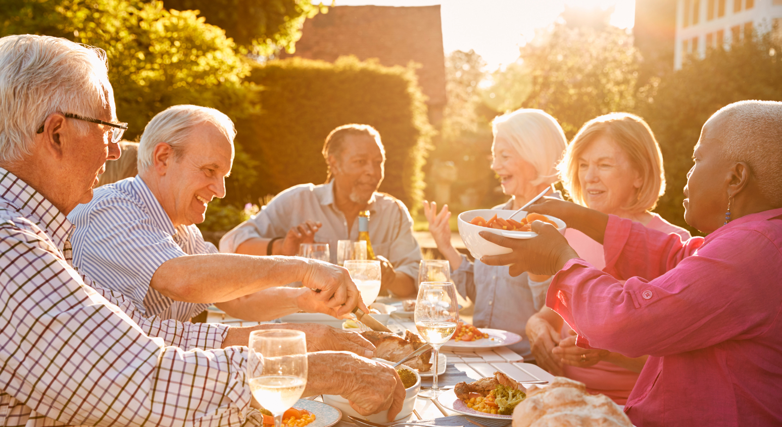 Six merry, beaming, attractive older adults sitting together joyfully sharing a meal on a lovely summer evening.